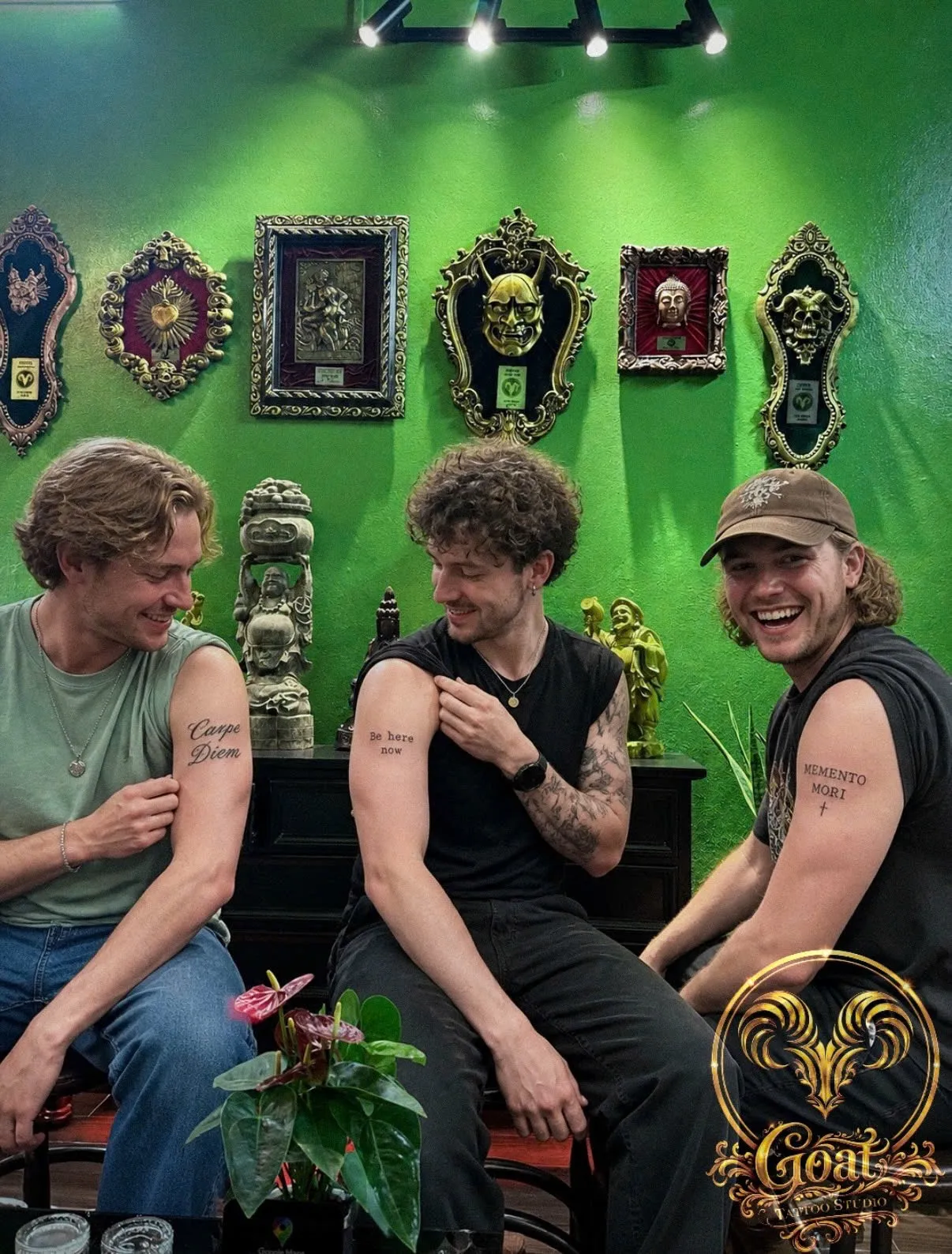 Three friends showing off matching minimalist lettering tattoos reading "Carpe Diem," "Be Here Now," and "Memento Mori" on their upper arms, done at GOAT Tattoo Studio in Hanoi's Old Quarter.