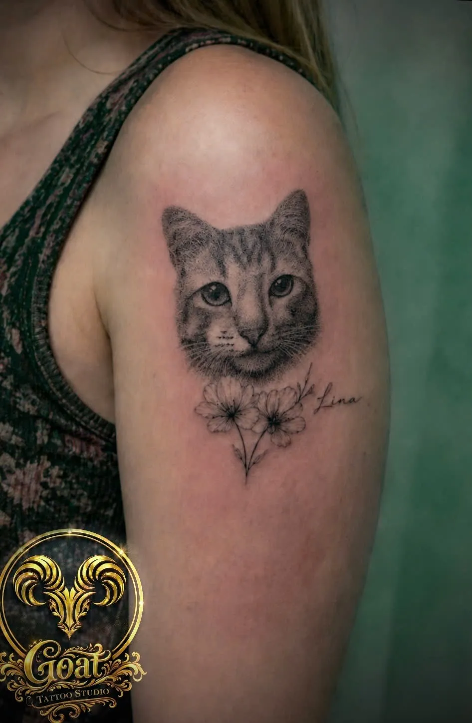 Black and grey realistic cat portrait named 'Lina' with fine line flowers on the upper arm, GOAT Tattoo Studio Hanoi.