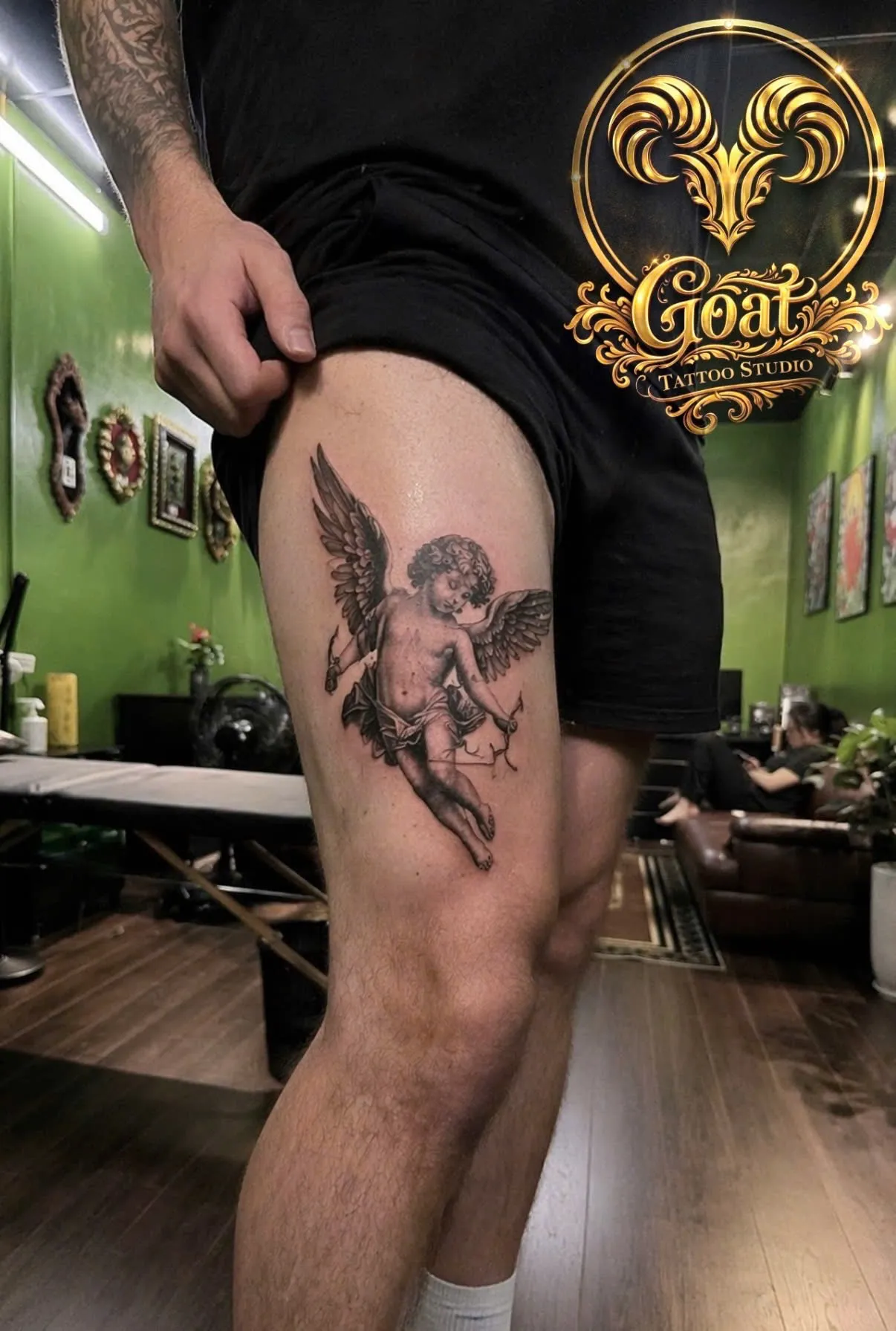 Black and grey cherub angel statue tattoo with bow and arrow on the thigh, by GOAT Tattoo Studio in Hanoi.