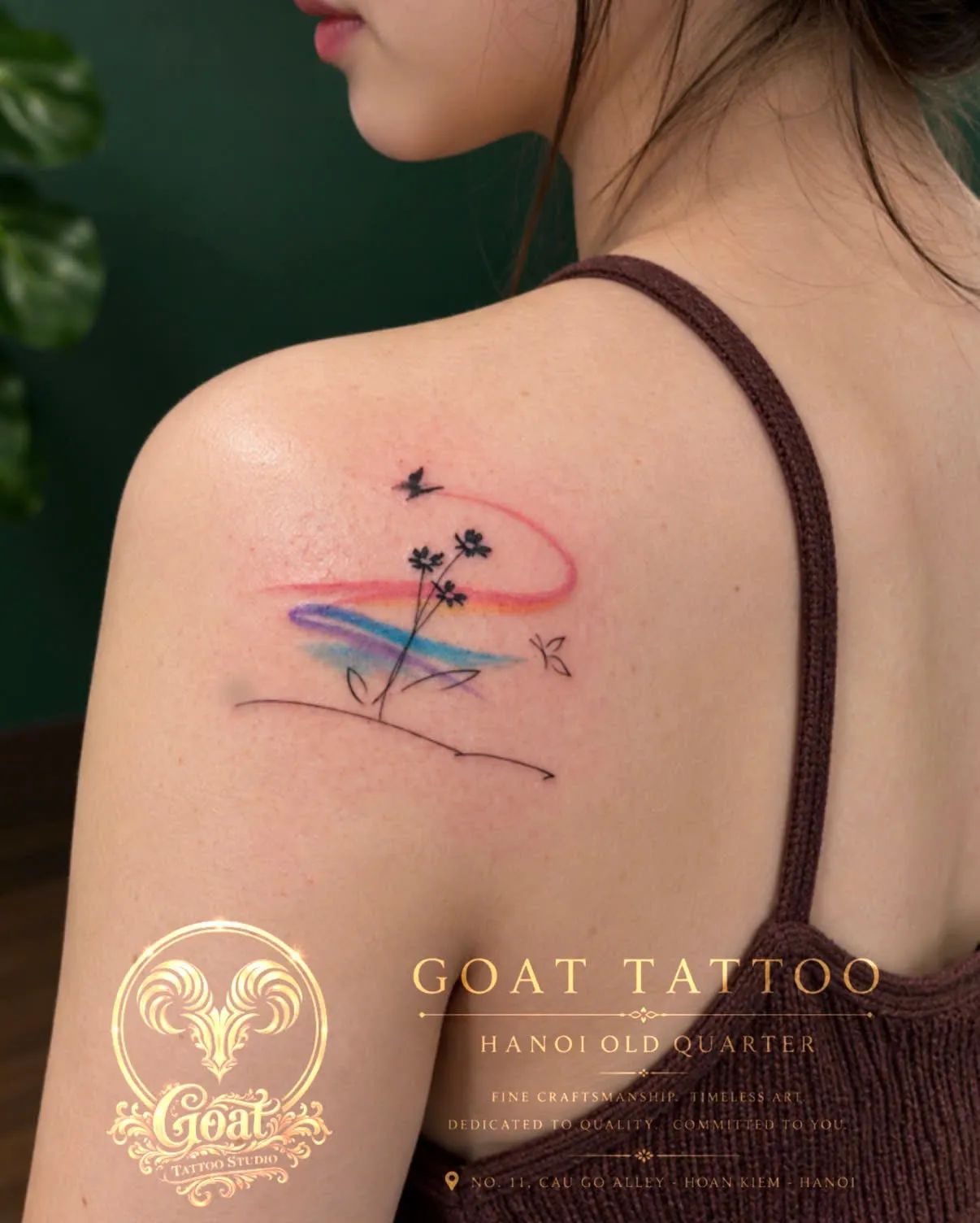A watercolor-style tattoo of a bird, wildflowers, and a rainbow swash on the shoulder blade, by GOAT Tattoo Studio in Hanoi's Old Quarter.