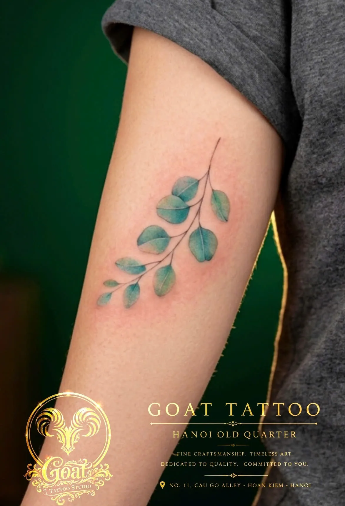 Watercolor-style eucalyptus branch tattoo in soft green and blue tones on the upper arm, GOAT Tattoo Studio Hanoi.