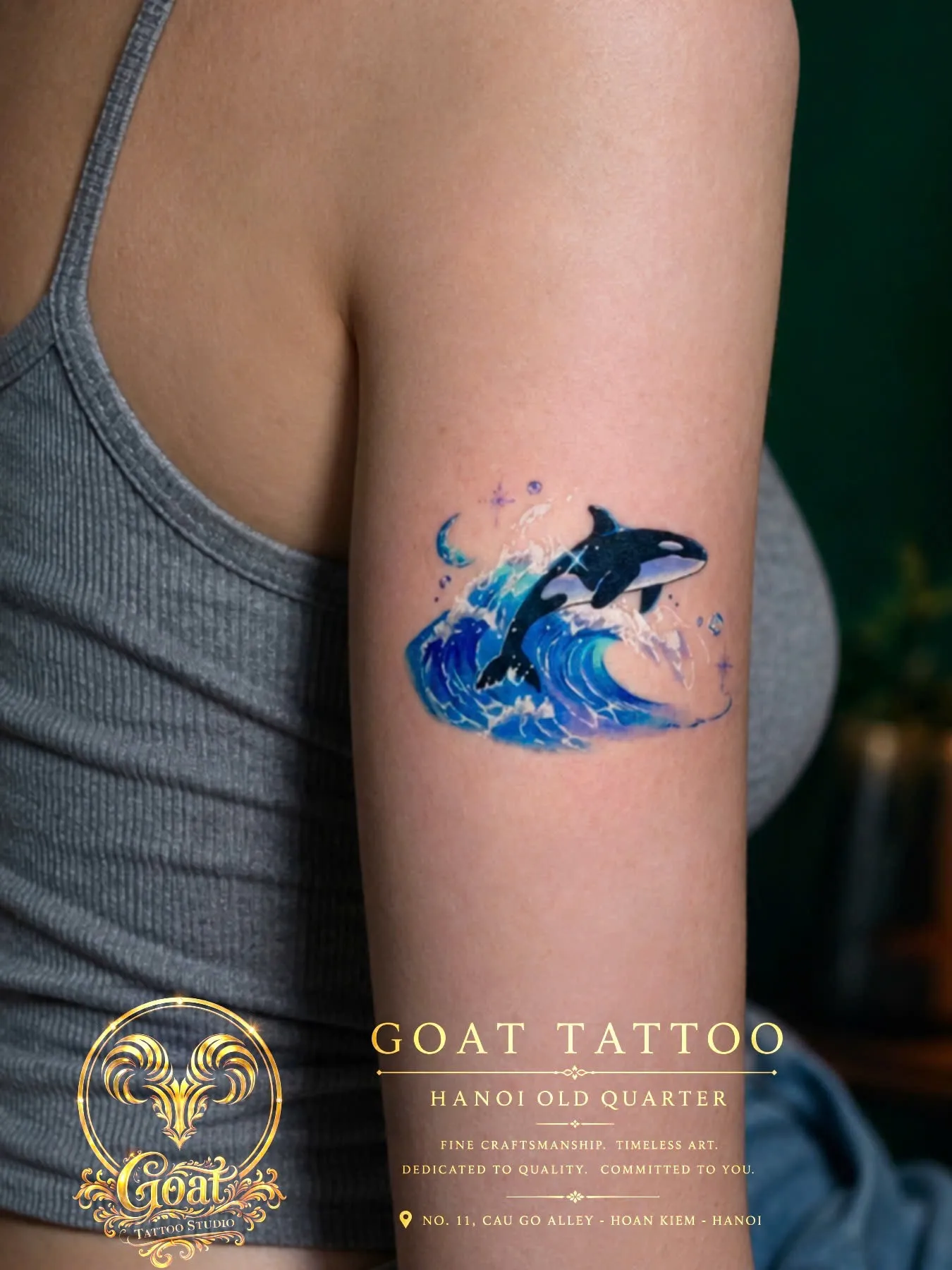 Colorful illustrative tattoo of an orca whale leaping through a crashing wave on the upper arm, by GOAT Tattoo Studio in Hanoi.