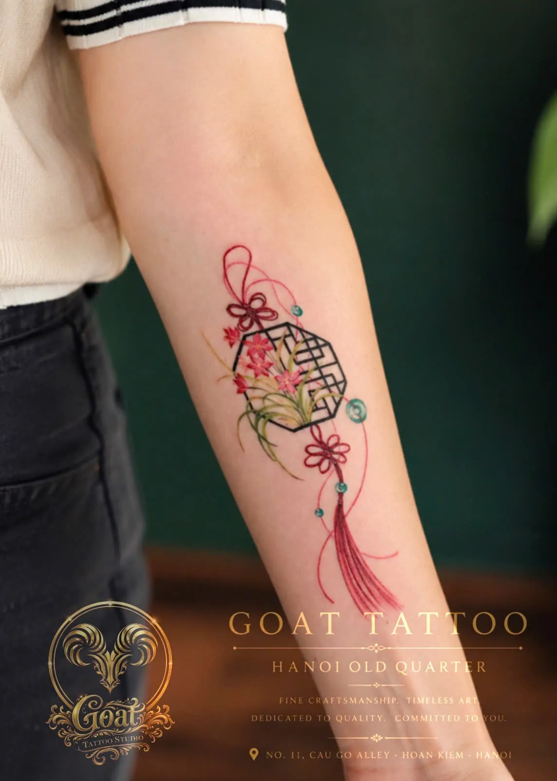 Colorful illustrative tattoo of a traditional ornamental knot with flowers and tassel on the forearm, by GOAT Tattoo Studio Hanoi.