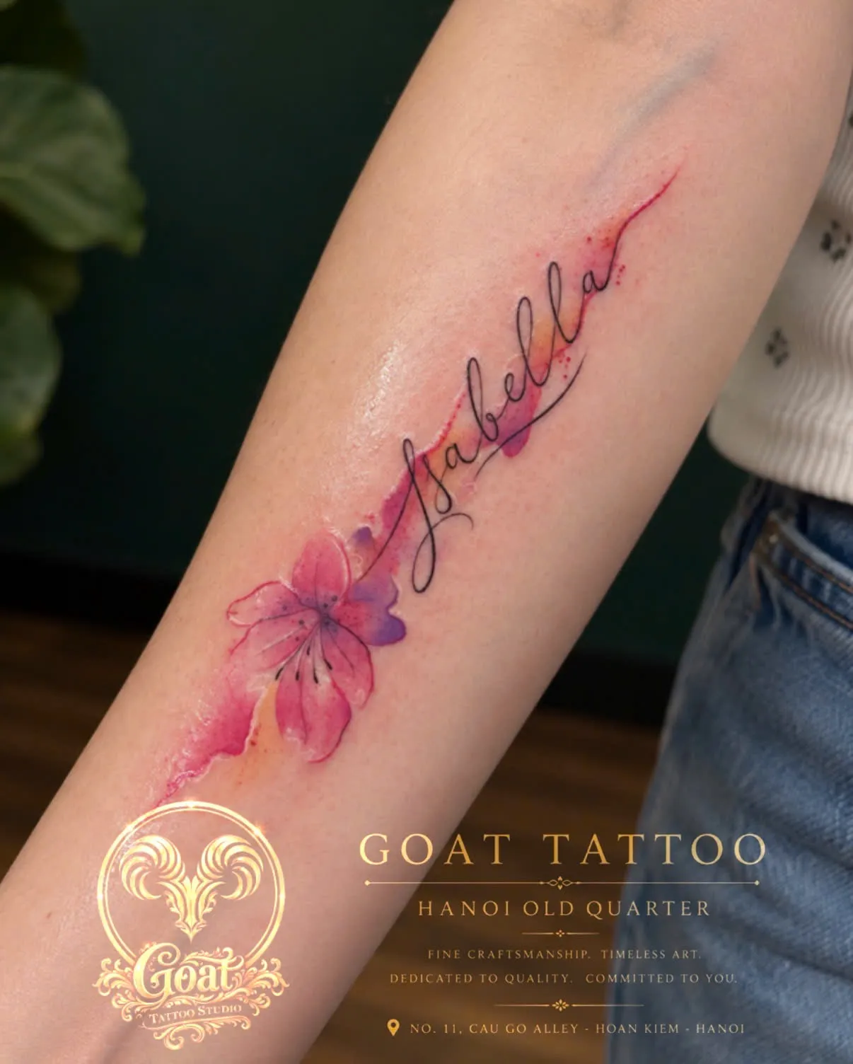 A cursive name tattoo flowing into a pink watercolor-style flower, on the forearm, by GOAT Tattoo Studio Hanoi.