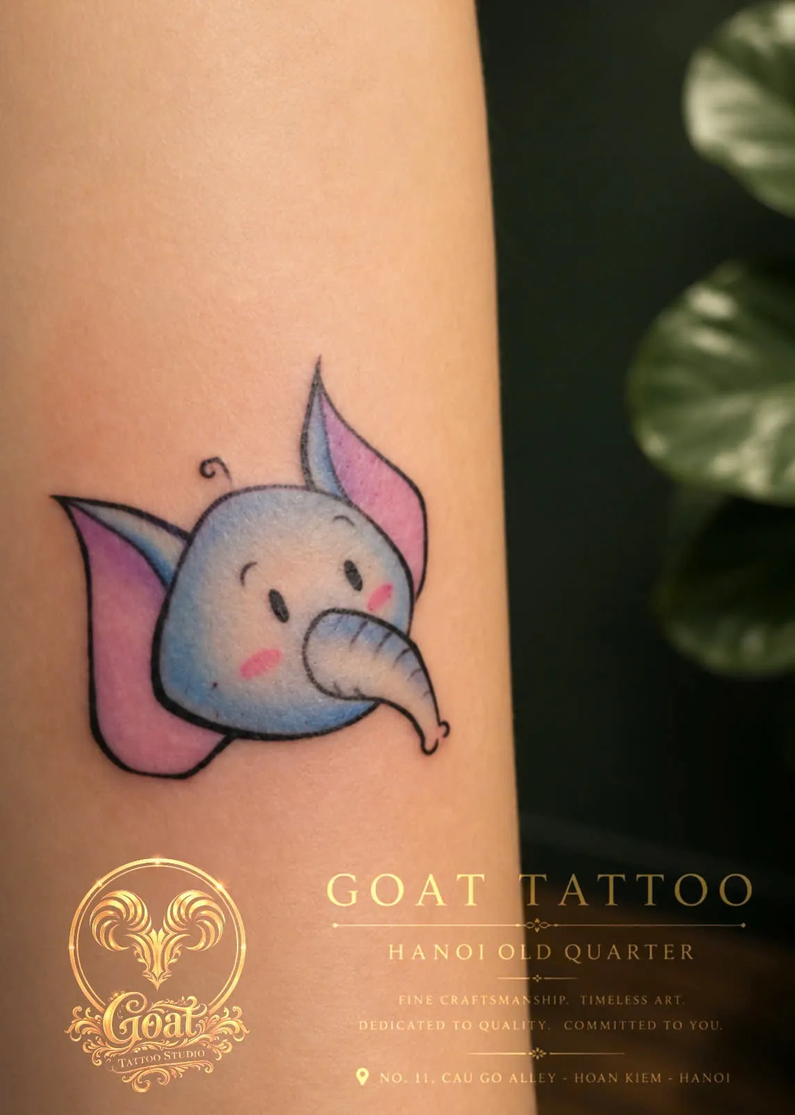 A playful color tattoo of a chubby blue elephant with pink ears and rosy cheeks on the forearm, done at GOAT Tattoo Studio in Hanoi's Old Quarter.