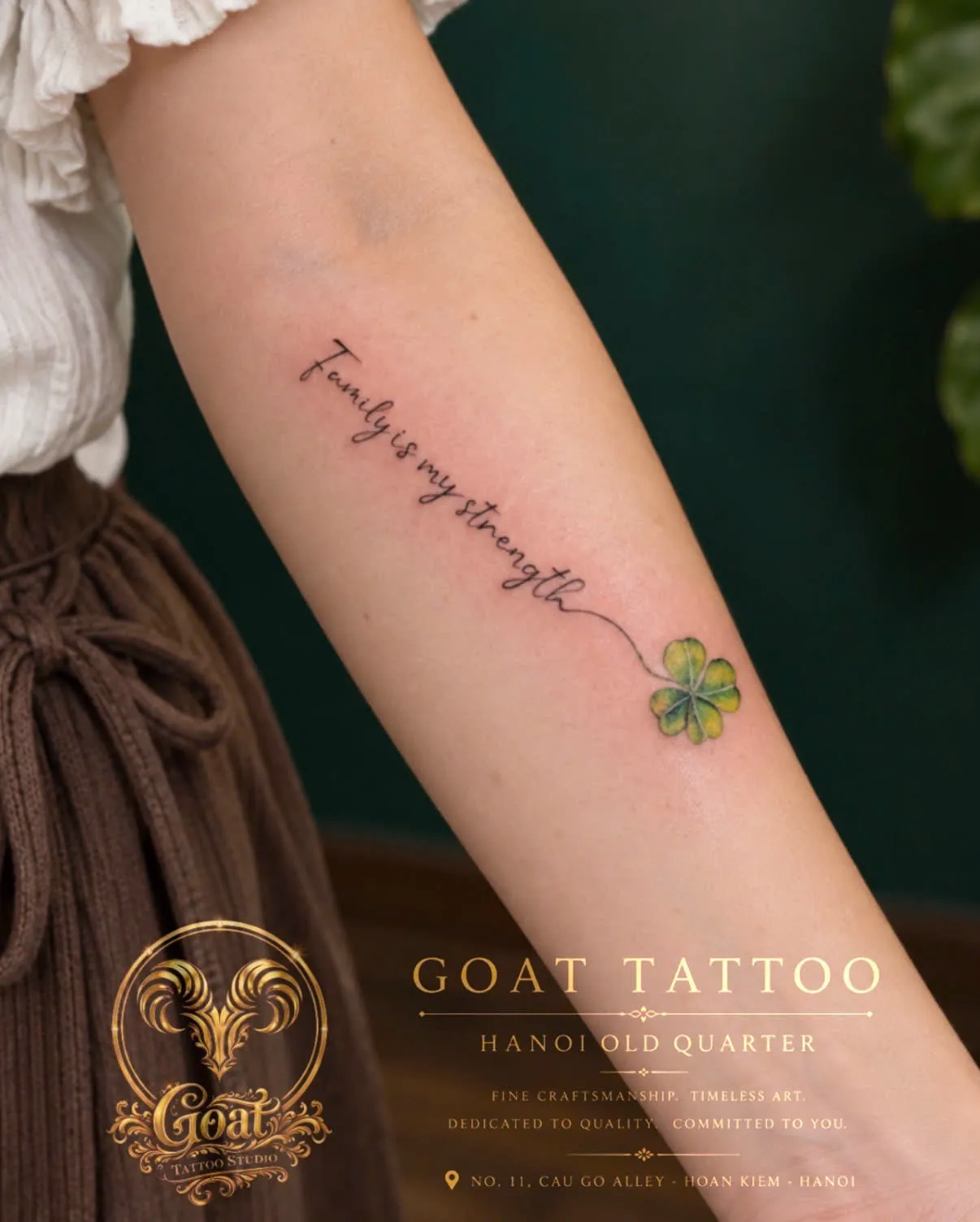 A cursive Family Is My Strength script paired with a four-leaf clover, tattooed on the forearm, by GOAT Tattoo Studio in Hanoi's Old Quarter.