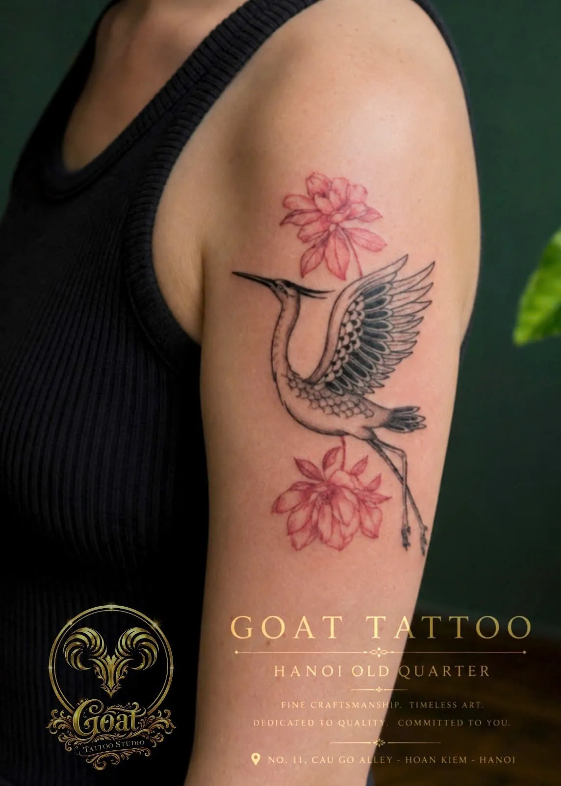 Fine line crane tattoo with delicate pink flower accents on the upper arm, by GOAT Tattoo Studio Hanoi.