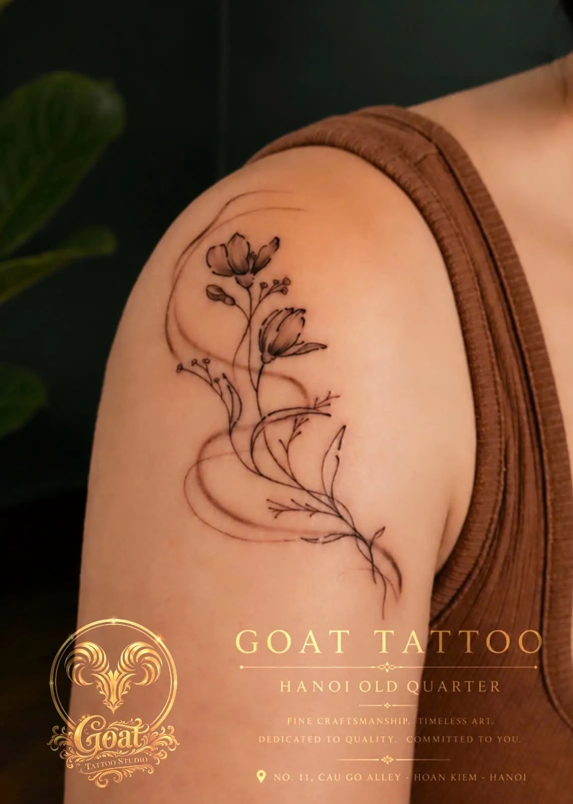 A delicate fine line tattoo of a flowering branch with flowing ribbon accents on the shoulder, done at GOAT Tattoo Studio in Hanoi's Old Quarter.