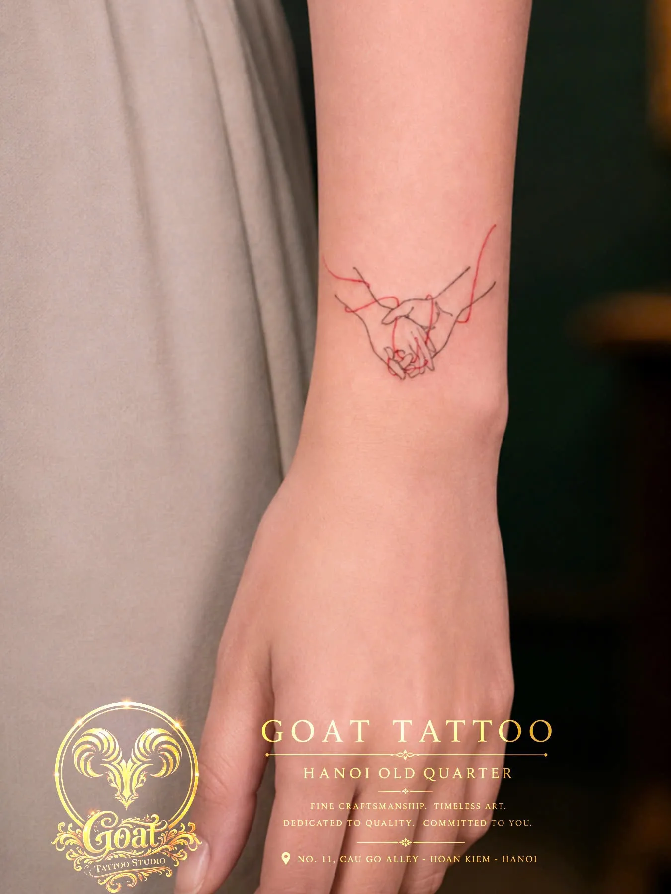Fine line tattoo of two hands holding with red thread accents wrapped around the wrist, by GOAT Tattoo Studio, Hanoi.
