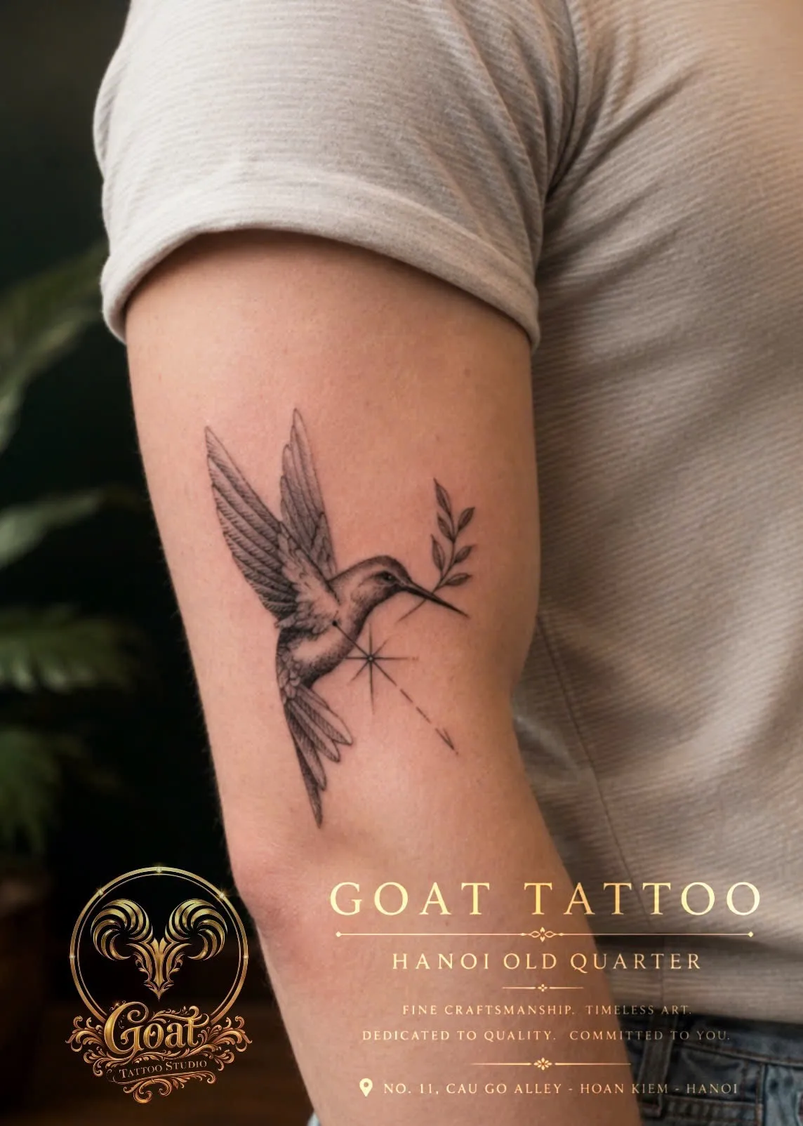 Delicate fine line hummingbird tattoo paired with a leafy sprig on the upper arm, GOAT Tattoo Studio Hanoi.