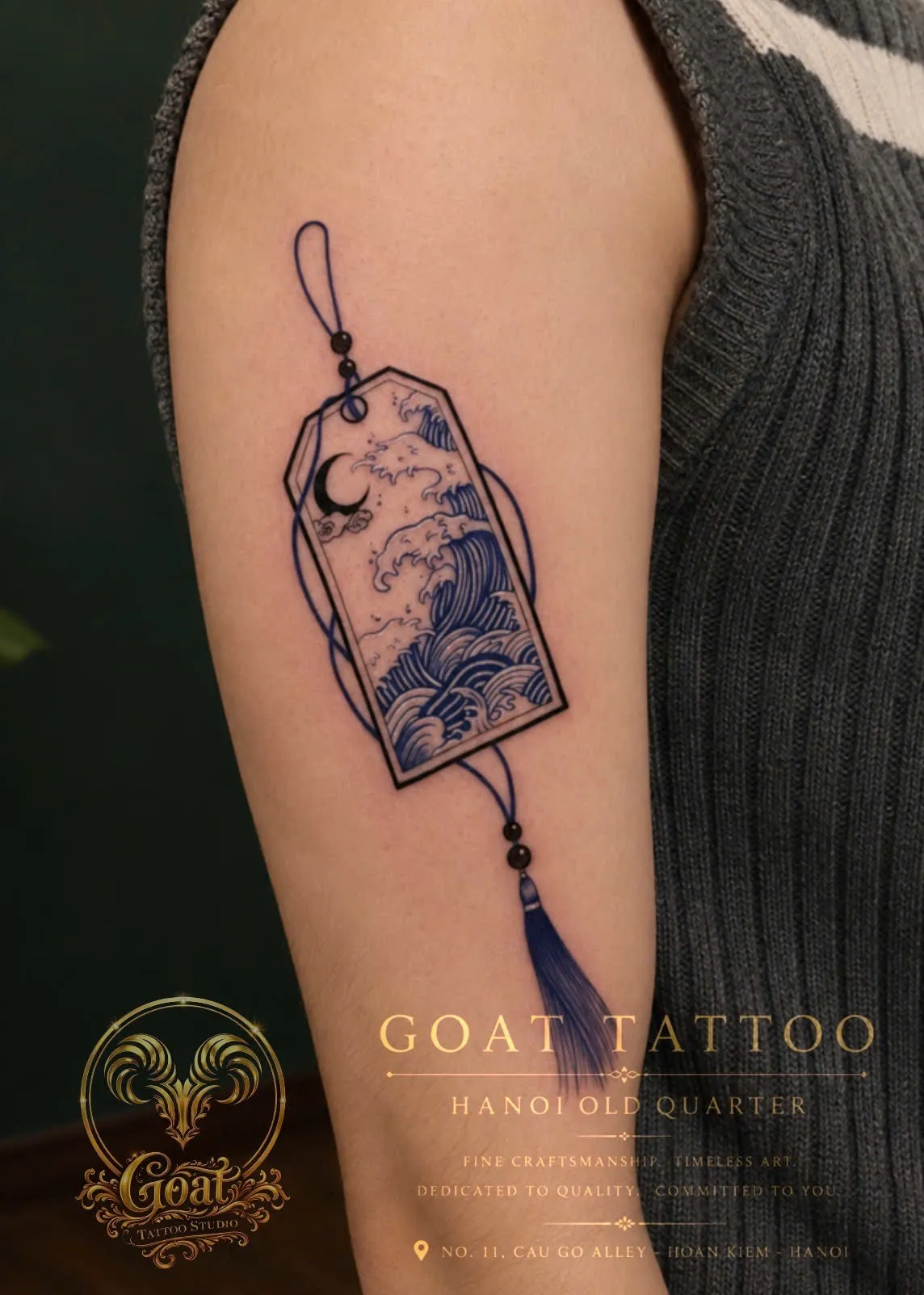 Fine line tattoo of an ornamental tag charm featuring ocean waves and a crescent moon, on the upper arm, by GOAT Tattoo Studio Hanoi.