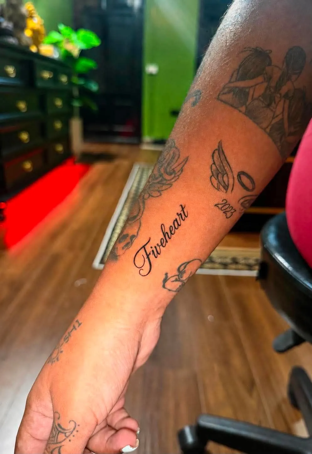 A black and grey forearm tattoo combining cursive lettering with angel wings, a halo, skulls, and a dated memorial detail, done at GOAT Tattoo Studio in Hanoi's Old Quarter.
