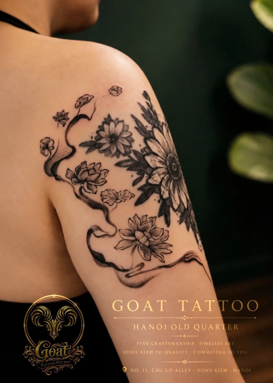 A detailed black and grey tattoo of a flowing lotus and daisy flower bouquet with ribbon trails on the shoulder, done at GOAT Tattoo Studio in Hanoi's Old Quarter.