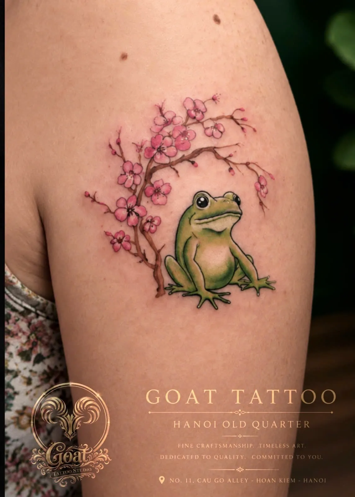 A charming color tattoo of a green frog seated beneath a blooming cherry blossom branch on the shoulder, done at GOAT Tattoo Studio in Hanoi's Old Quarter.