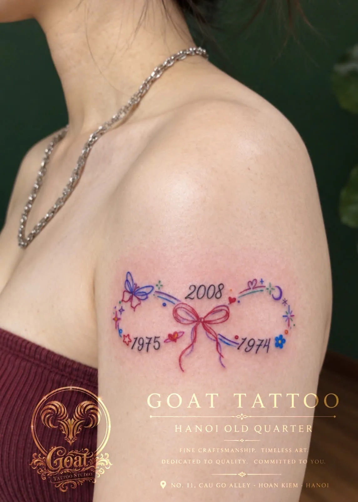 A colorful infinity symbol tattoo with family birth years, a butterfly, and a bow, on the upper arm, by GOAT Tattoo Studio Hanoi.