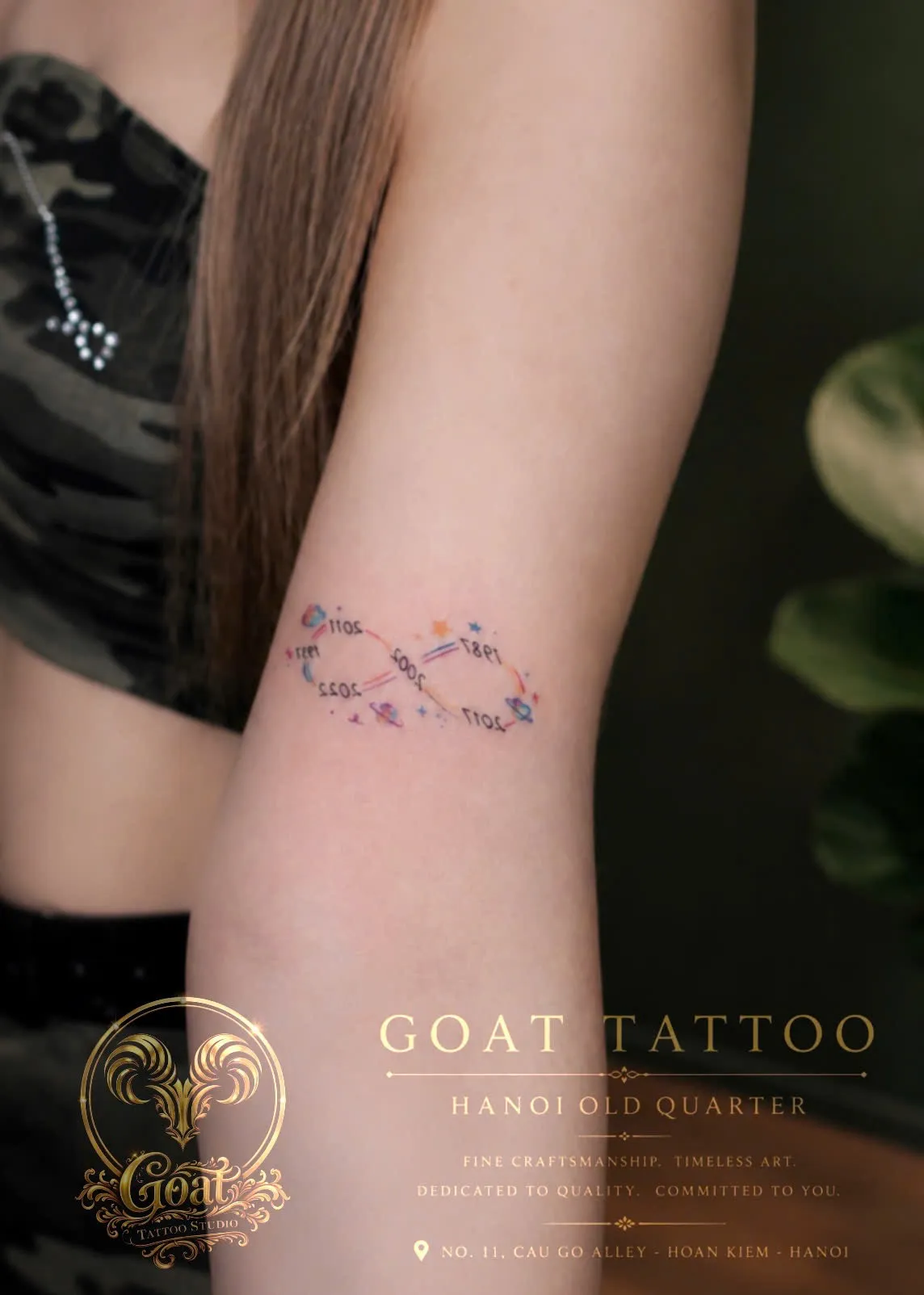 A delicate fine-line infinity symbol tattoo entwined with tiny colorful stars, planets, and meaningful family dates on the upper arm, done at GOAT Tattoo Studio in Hanoi's Old Quarter.