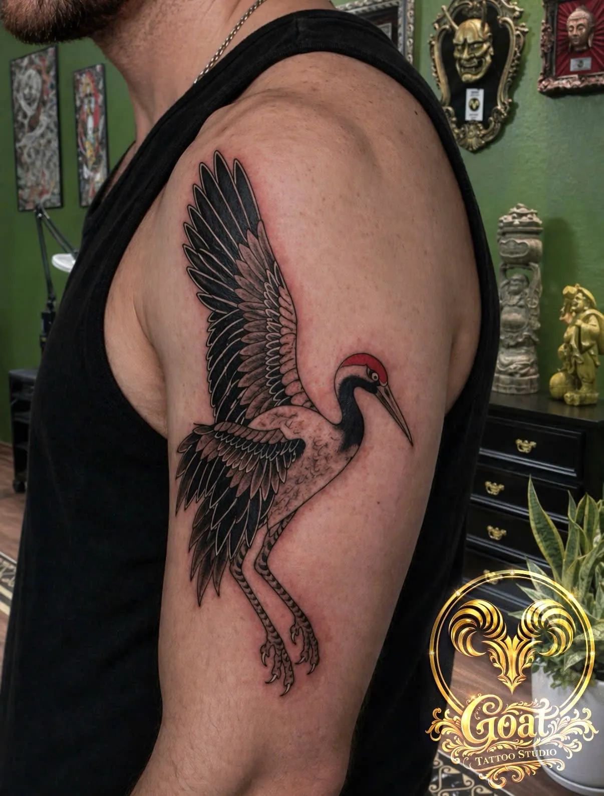 Black and grey crane tattoo with a red crown accent, wings spread across the upper arm, by GOAT Tattoo Studio Hanoi.