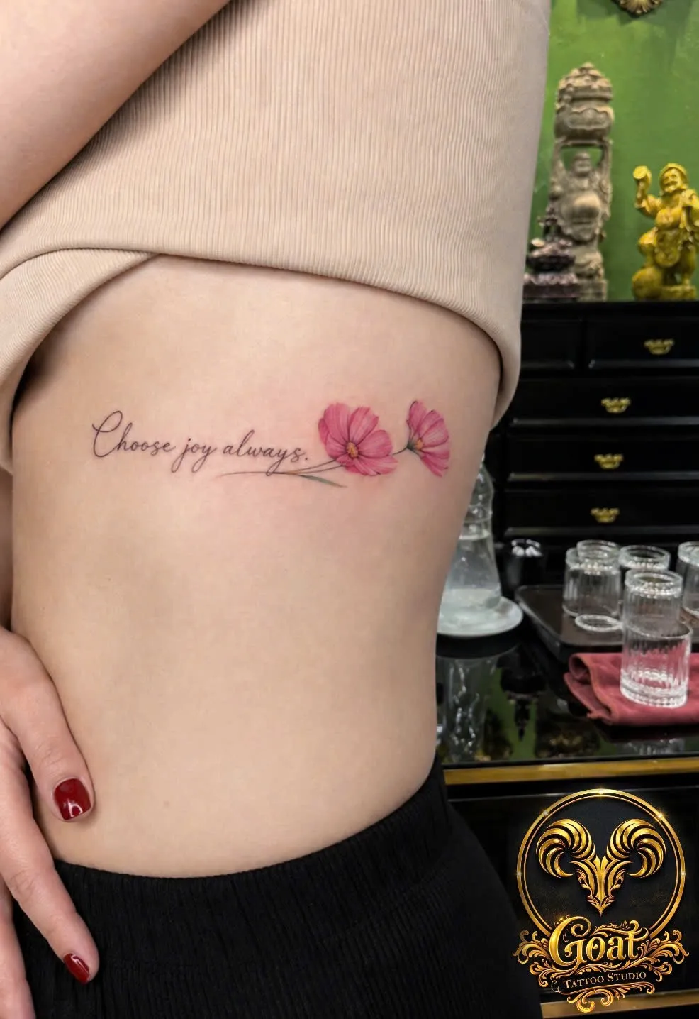 Elegant cursive lettering 'Choose joy always' with small pink flowers on the ribs, by GOAT Tattoo Studio in Hanoi.