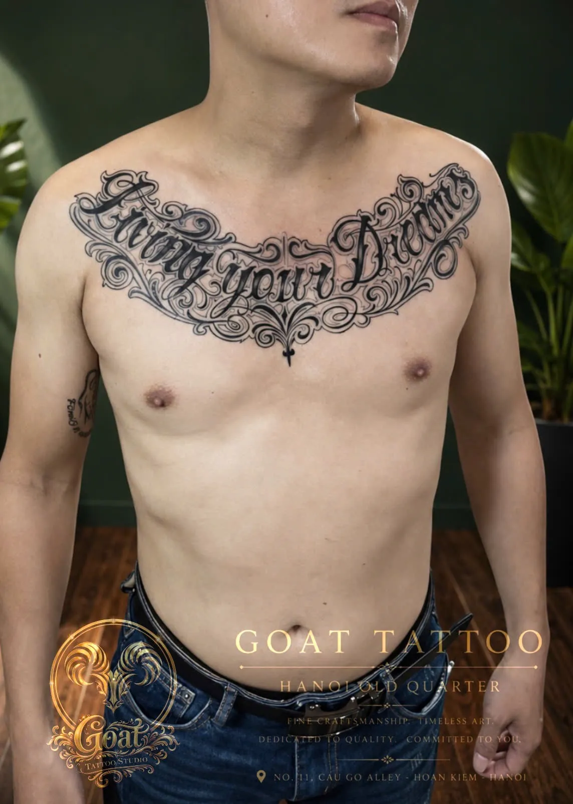 Ornate script lettering tattoo reading 'Living your Dreams' across the chest, by GOAT Tattoo Studio Hanoi.