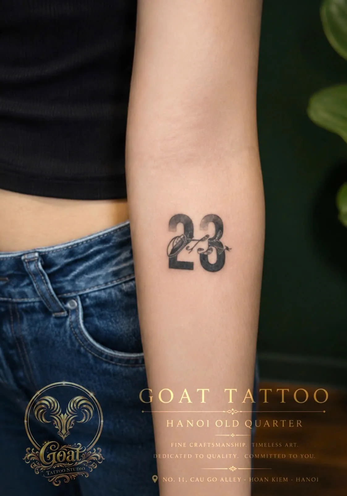 A small bold numeral tattoo of "23" accented with a delicate smoking pipe design on the forearm, done at GOAT Tattoo Studio in Hanoi's Old Quarter.