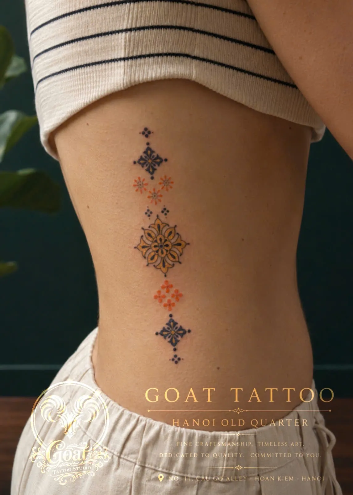 A chain of colorful ornamental medallions in blue, gold, and orange down the ribs, by GOAT Tattoo Studio Hanoi.
