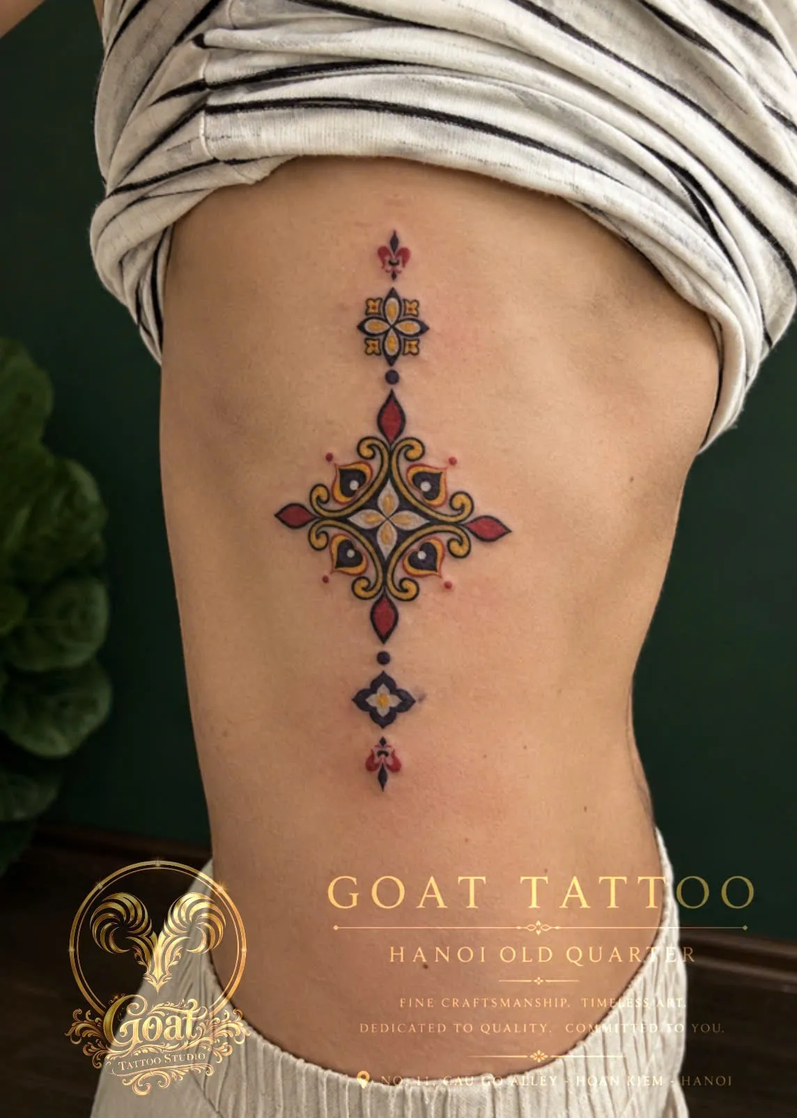 A colorful ornamental medallion tattoo with gold, red, and black detailing down the ribs, by GOAT Tattoo Studio Hanoi.