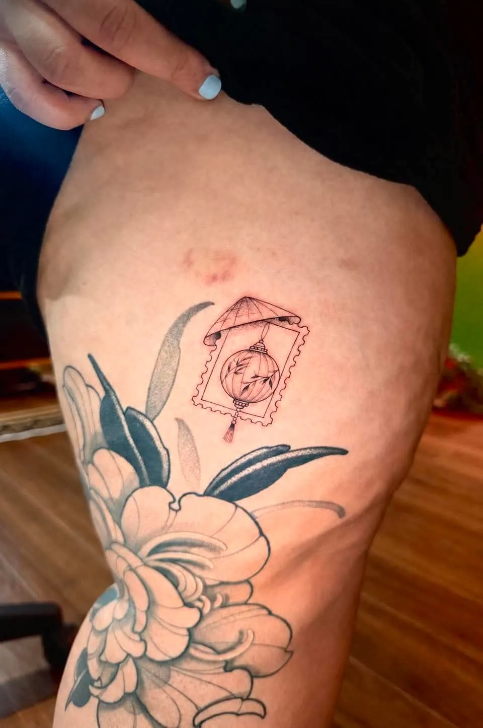 A fine-line hip tattoo pairing a large peony flower with a Vietnamese conical-hat lantern styled as a vintage postage stamp, done at GOAT Tattoo Studio in Hanoi's Old Quarter.