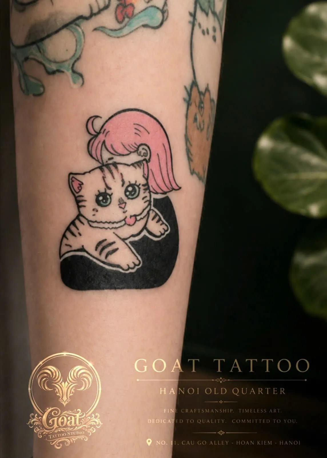A whimsical color tattoo of a wide-eyed kitten with pink hair peeking from a dark silhouette on the forearm, done at GOAT Tattoo Studio in Hanoi's Old Quarter.