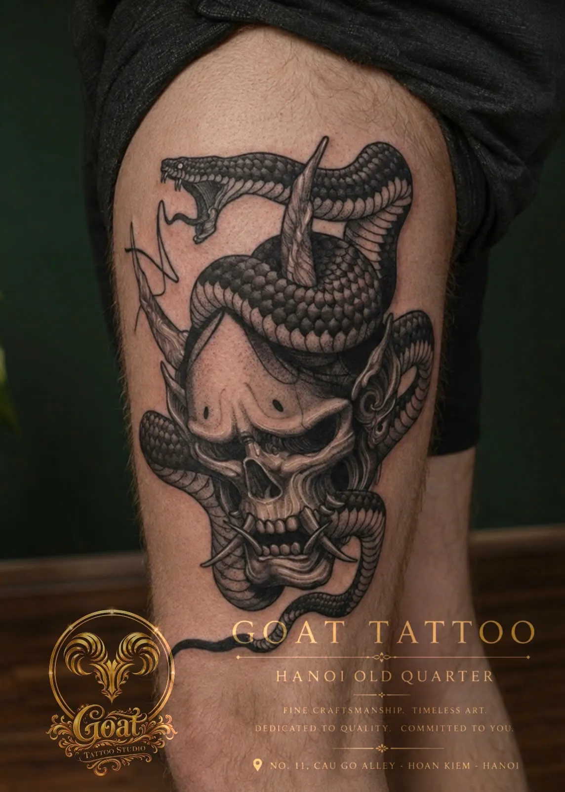A black and grey skull mask wrapped in a coiling snake, tattooed on the thigh, by GOAT Tattoo Studio Hanoi.