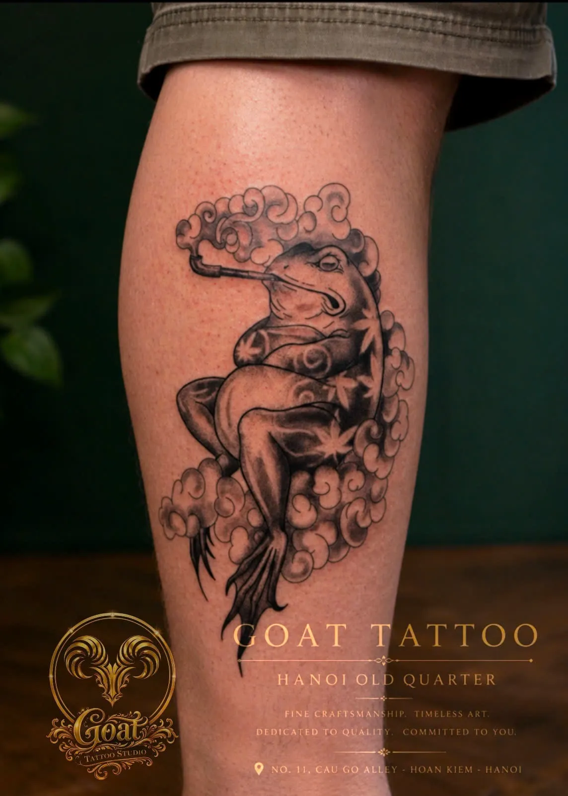 A black and grey illustrative toad smoking a pipe amid swirling clouds, on the calf, by GOAT Tattoo Studio in Hanoi's Old Quarter.