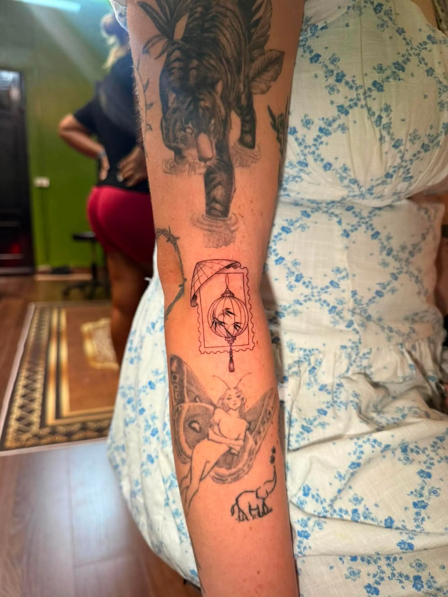 A fine-line forearm sleeve in progress featuring a tiger on a pedestal, a Vietnamese lantern styled as a postage stamp, a winged fairy, and a small elephant, done at GOAT Tattoo Studio in Hanoi's Old Quarter.
