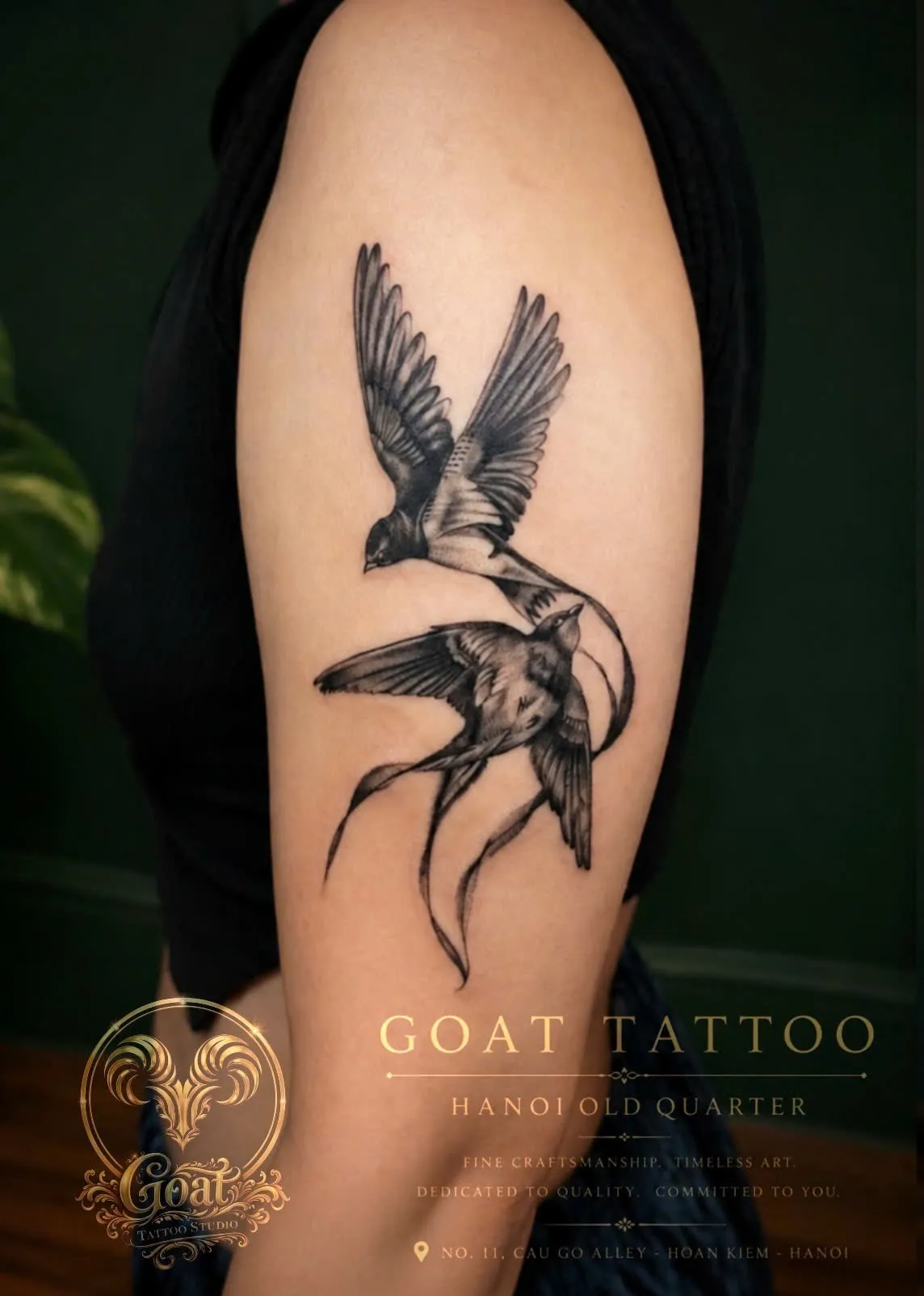 A detailed black and grey tattoo of two swallows in flight trailing long ribbon-like tails on the upper arm, done at GOAT Tattoo Studio in Hanoi's Old Quarter.