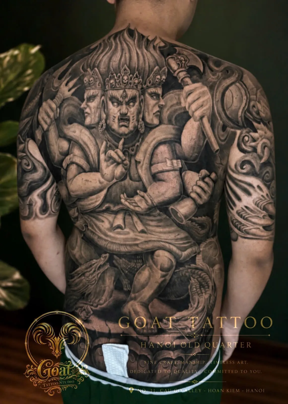 A vivid black and grey full back tattoo of a fierce three-faced guardian deity wielding a mace, done at GOAT Tattoo Studio in Hanoi's Old Quarter.